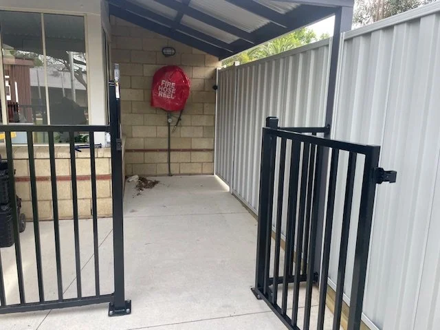 Outdoor area with a white fence, black gate, and a red fire hose reel mounted on a brick wall under a metal roof.