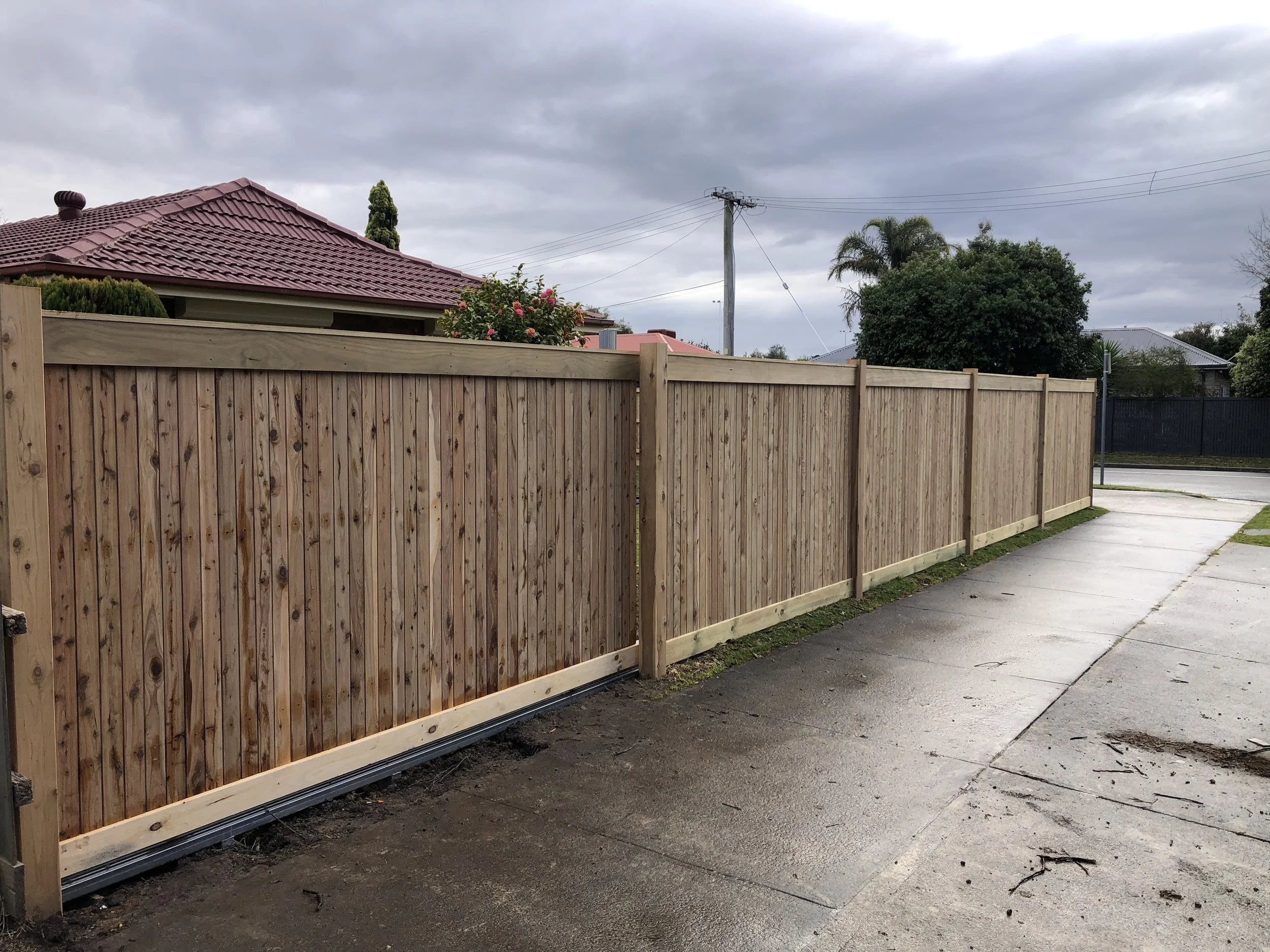 New wooden fence along a sidewalk next to residential houses with cloudy sky overhead.