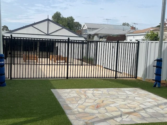 Backyard with artificial grass, a stone patio, and a black metal fence with wooden benches inside.