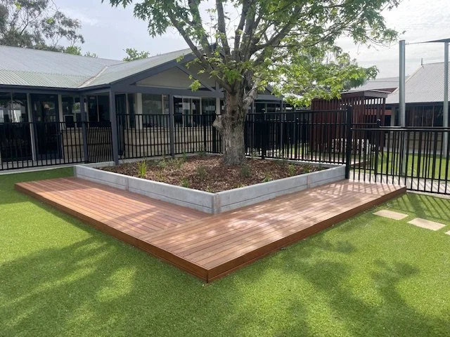 A backyard with a raised garden bed surrounding a tree, a wooden deck, and a black metal fence, with a house and a trampoline in the background.
