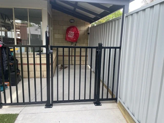 Outdoor backyard area with a black gate, a brick wall, a fire hose reel, and a covered patio with a sloped roof.