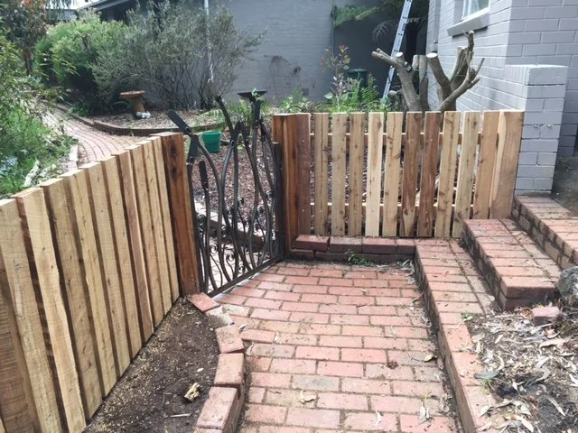 A brick pathway with a wooden fence on either side and a metal gate at the end. There are plants and garden elements in the background.