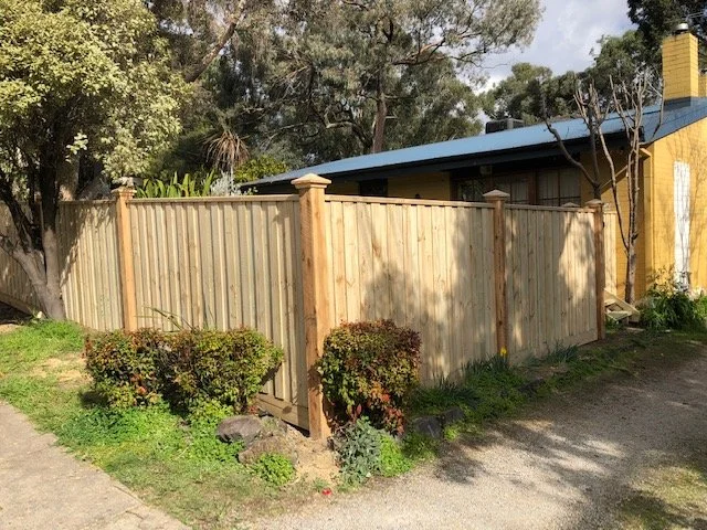 Wooden privacy fence with bushes and trees nearby, yellow building with chimney in background.