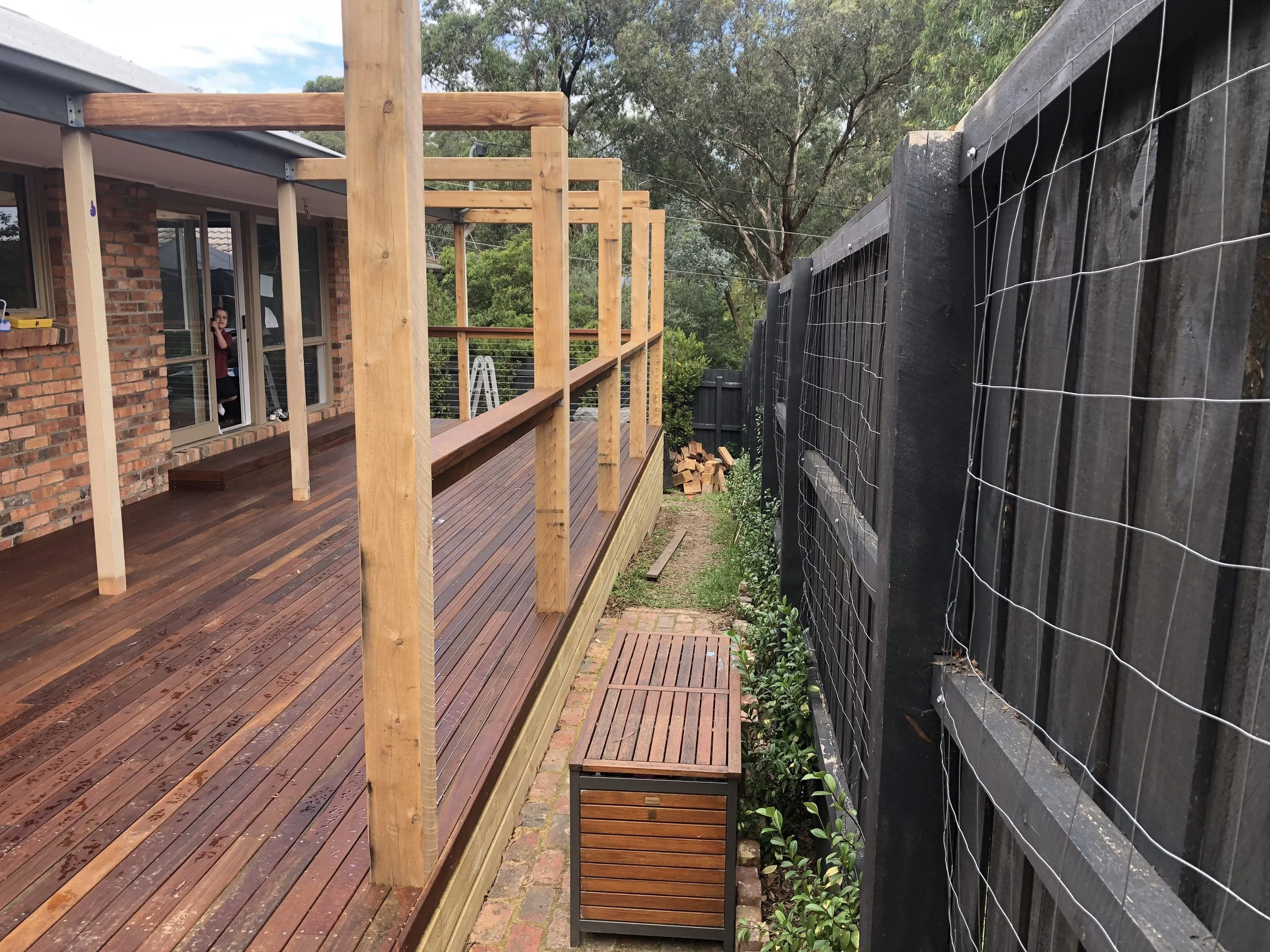 A backyard deck under construction with vertical wooden posts, a brick house with windows, and a black wooden fence with wire mesh on top.