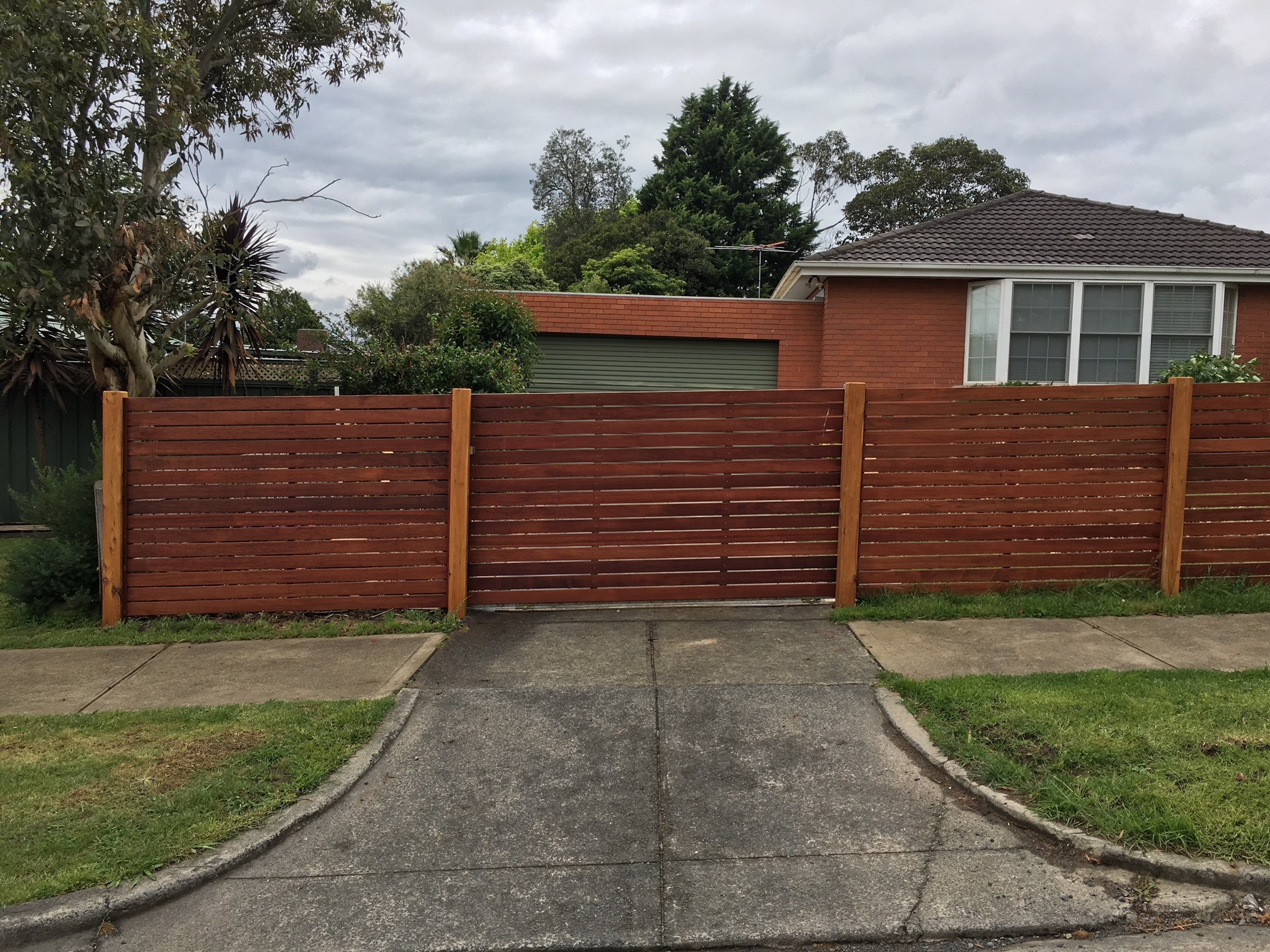 A wooden fence with a gate spans across a sidewalk and a driveway in front of a brick house with a bay window. There are trees and shrubs in the background under a cloudy sky.