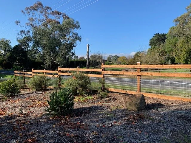 A landscaped area with a wooden fence, small bushes, and a large rock, with a road and trees in the background under a clear blue sky.