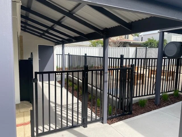 Fenced outdoor patio area with a black metal gate, wooden benches, and a covered roof.