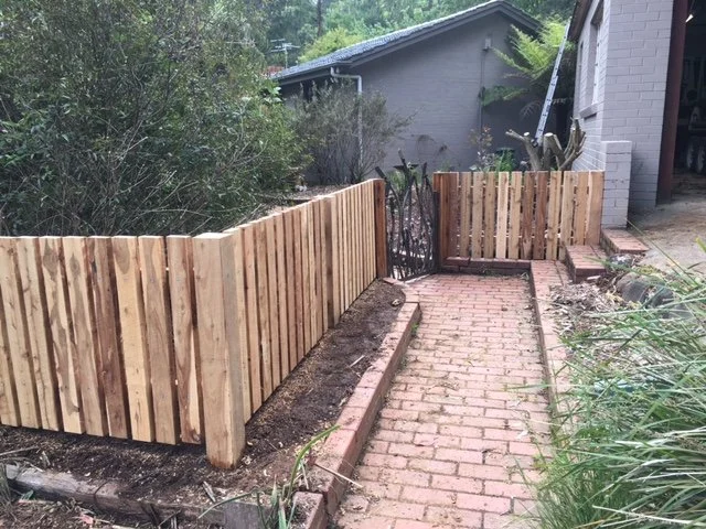 Newly installed wooden fence along a brick sidewalk outside a house, with some plants and bushes nearby.