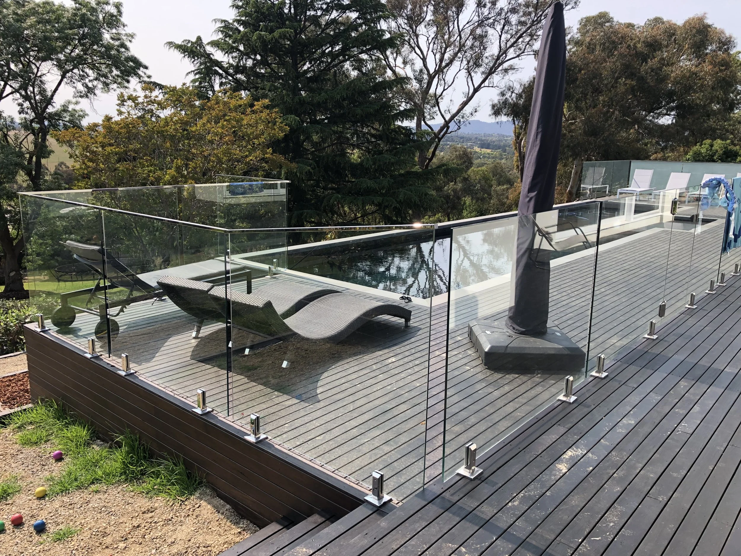 Outdoor deck with lounge chairs, a glass safety barrier, and a pool, surrounded by trees and distant landscape.