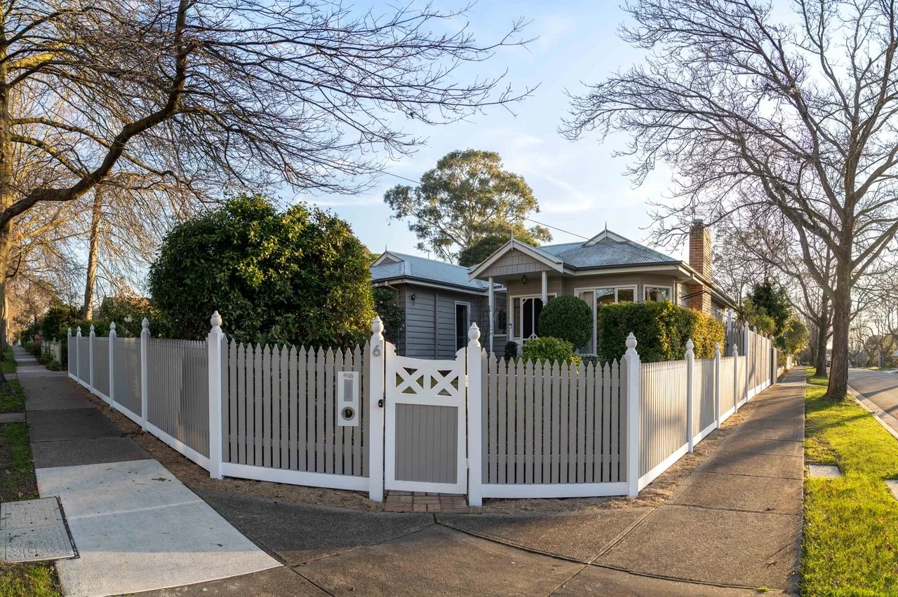 A house with a gray exterior, white picket fence, and landscaped bushes, trees, and a sidewalk in front.