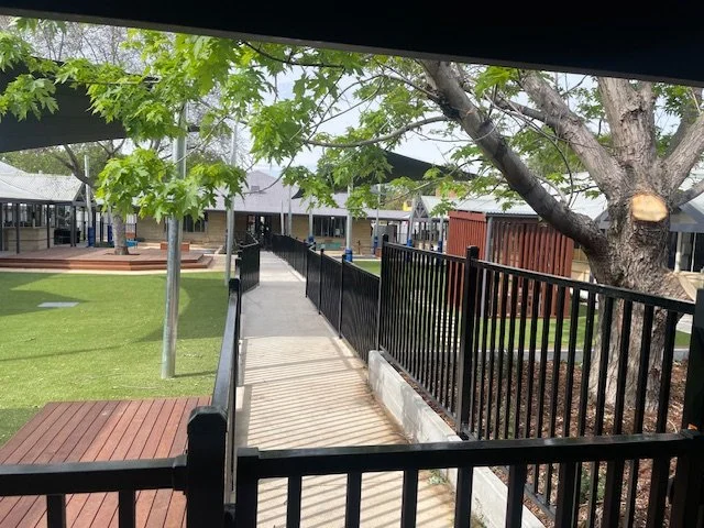 A walkway with a black metal fence on both sides, shaded by green trees, leading to a building in the distance, with a grassy area on the left and a large tree on the right.