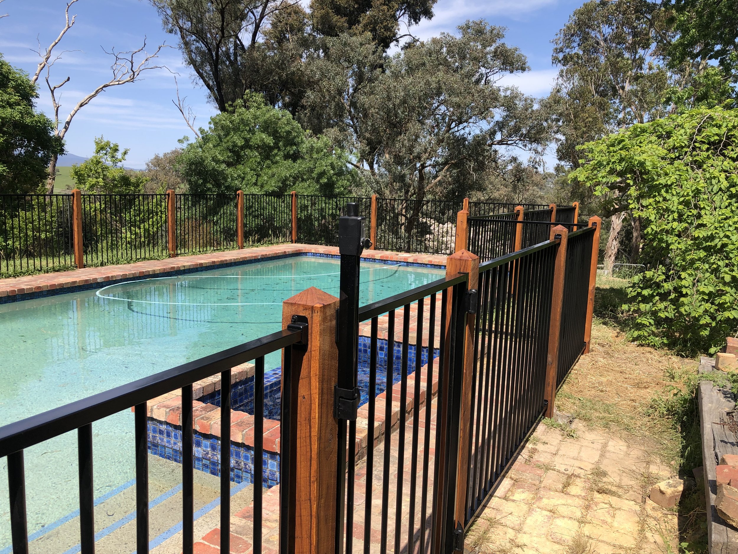 A fenced backyard pool with blue and brick edges, surrounded by trees and greenery under a blue sky.
