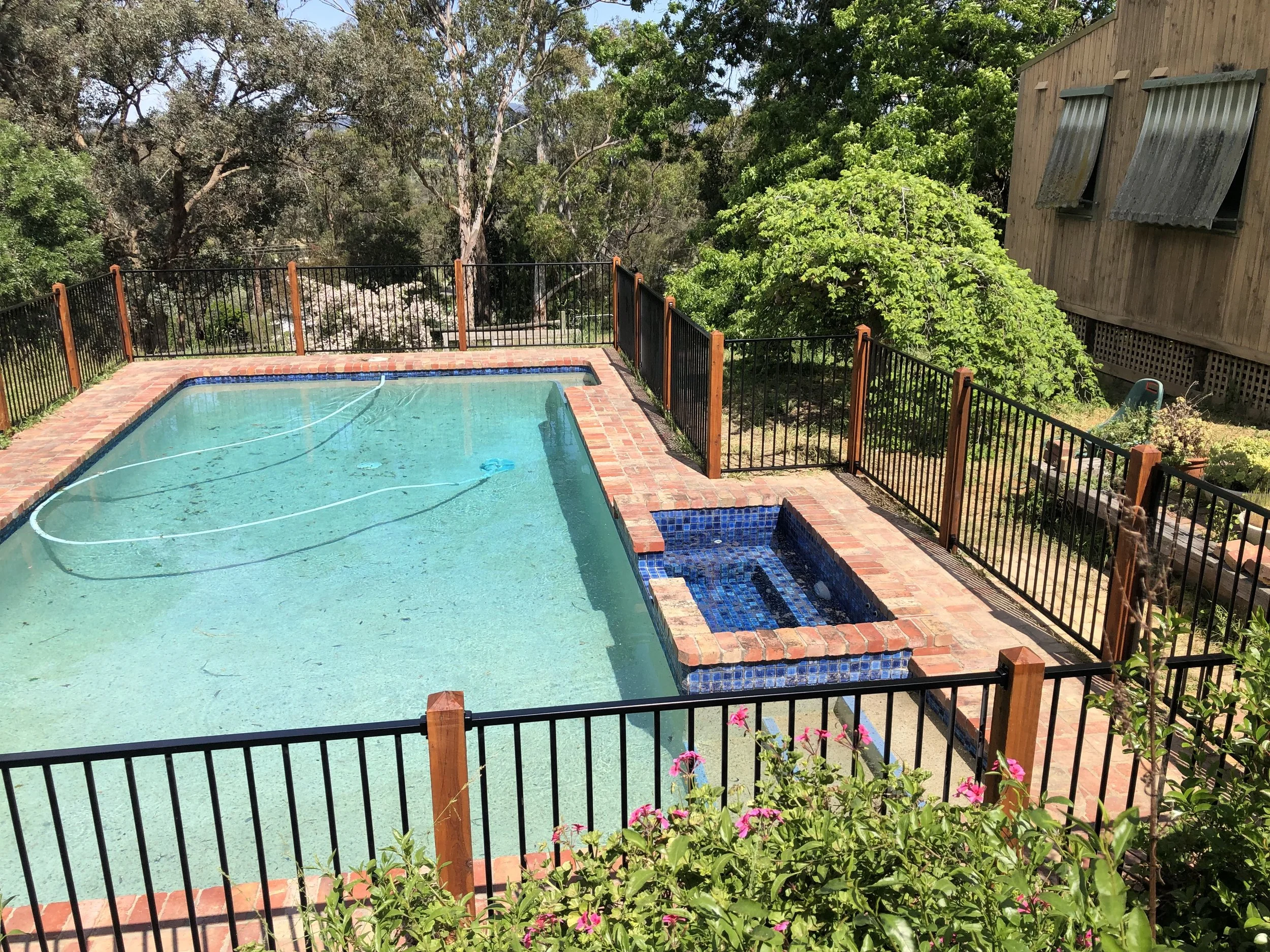 Residential backyard swimming pool with black metal fence, brick border, and hot tub, surrounded by trees and a wooden house.