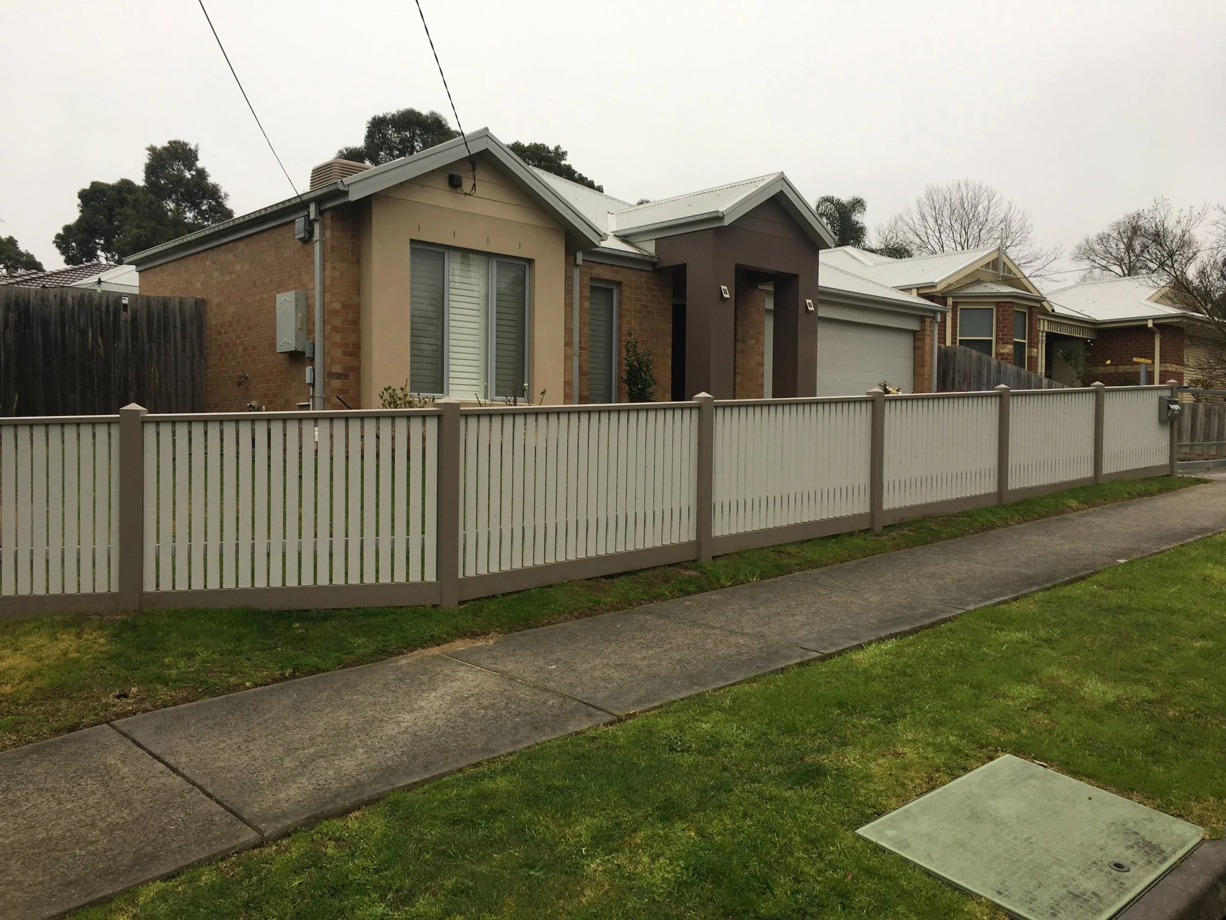 Front view of a modern single-story house with a white picket fence and sidewalk in front. The house has beige and brown exterior walls, large windows with white shutters, and a grey roof. There are some small plants by the entrance and a grassy lawn