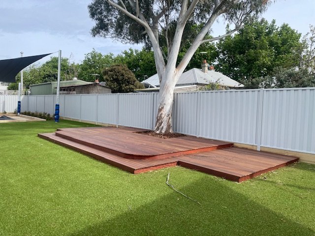 Backyard with a wooden deck built around a large tree, a green grassy lawn, a white fence, and trees in the background.