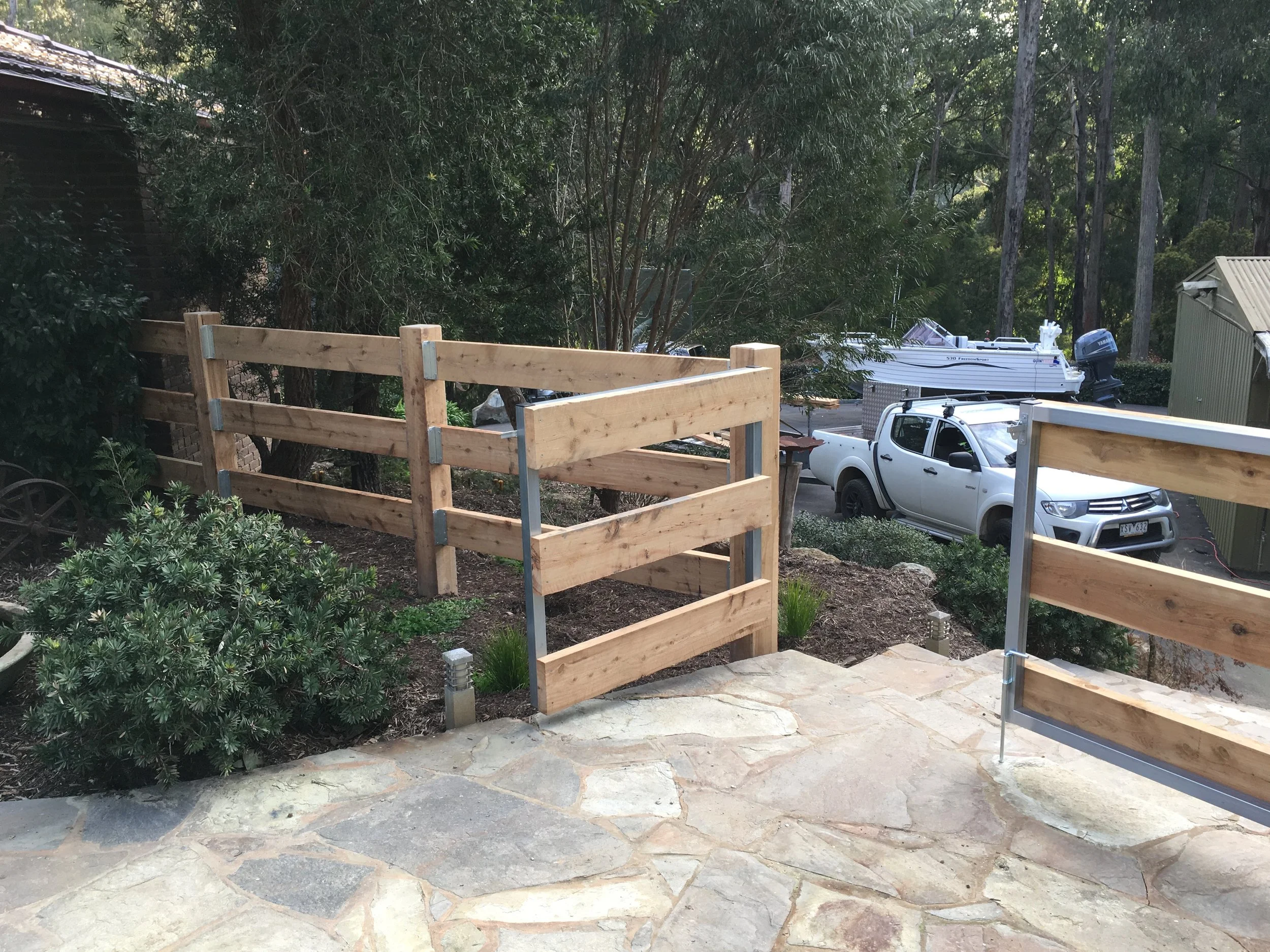 Partially constructed wooden fence surrounding a landscaped area with shrubs, adjacent to a stone patio, with a driveway and parked vehicles in the background.