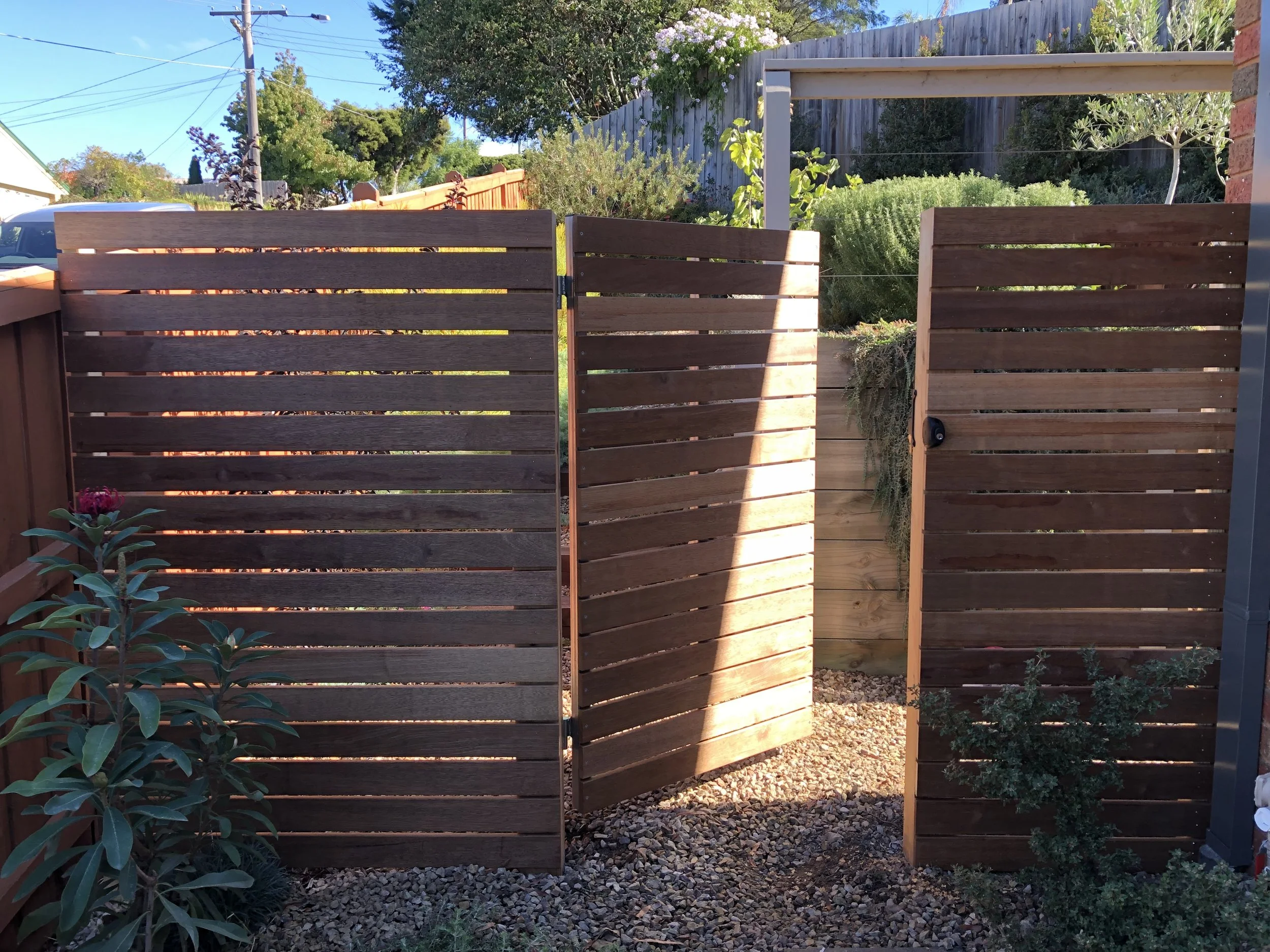 A wooden privacy outdoor shower area with a small door, surrounded by plants and garden landscaping, in a backyard.