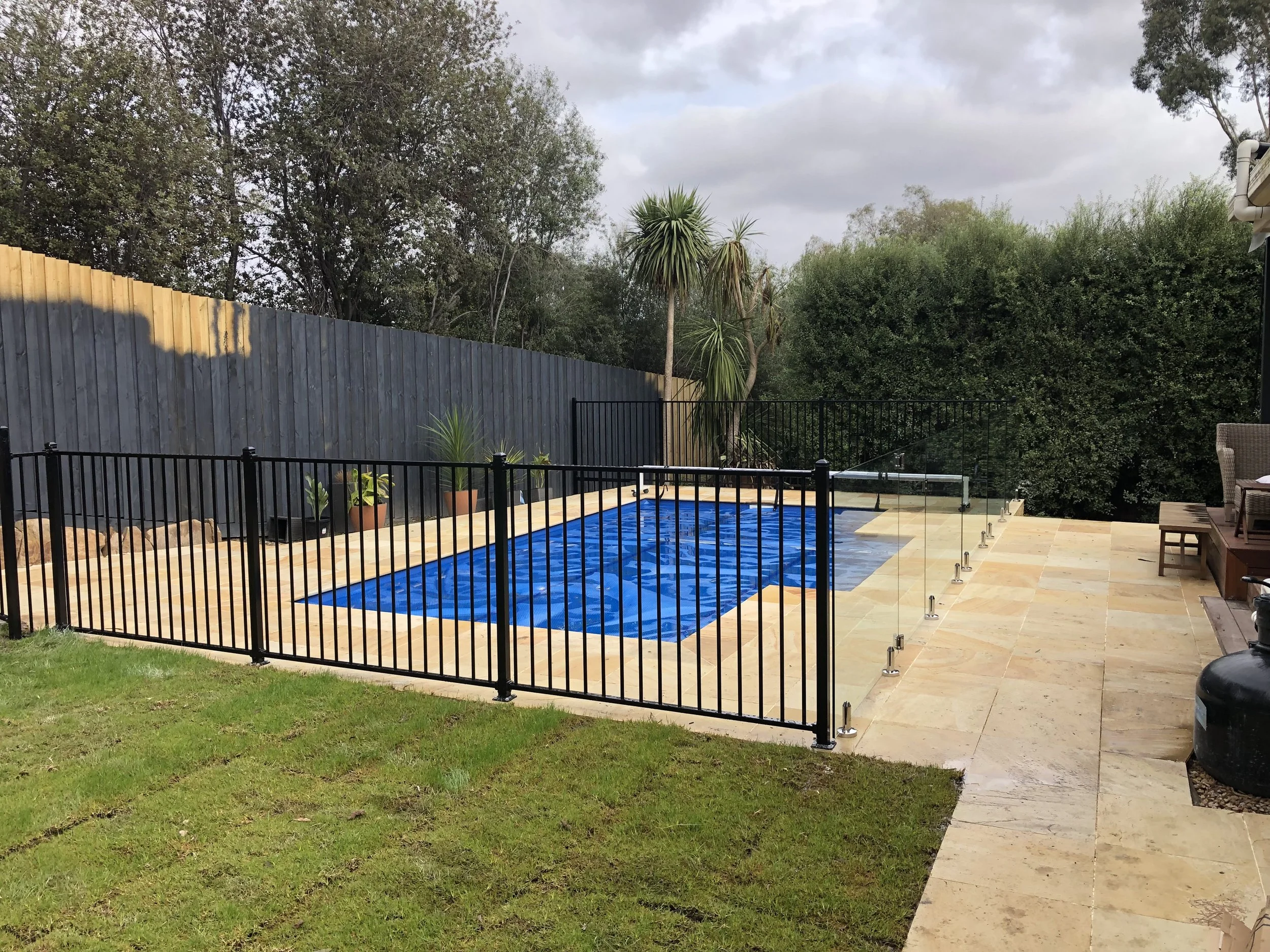 Backyard with in-ground swimming pool with a black safety fence, surrounded by beige stone paving, with potted plants, and lush trees in the background.