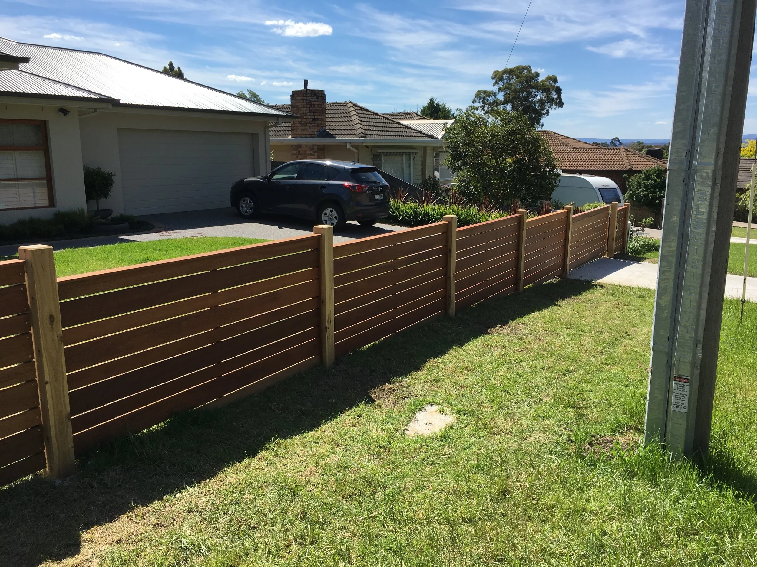 View of a suburban front yard and driveway with a new wooden fence, a black SUV, and neighboring houses under blue sky.