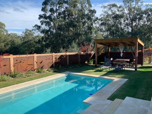Backyard with a swimming pool, a covered seating area with outdoor furniture, and a wooden fence with trees in the background.