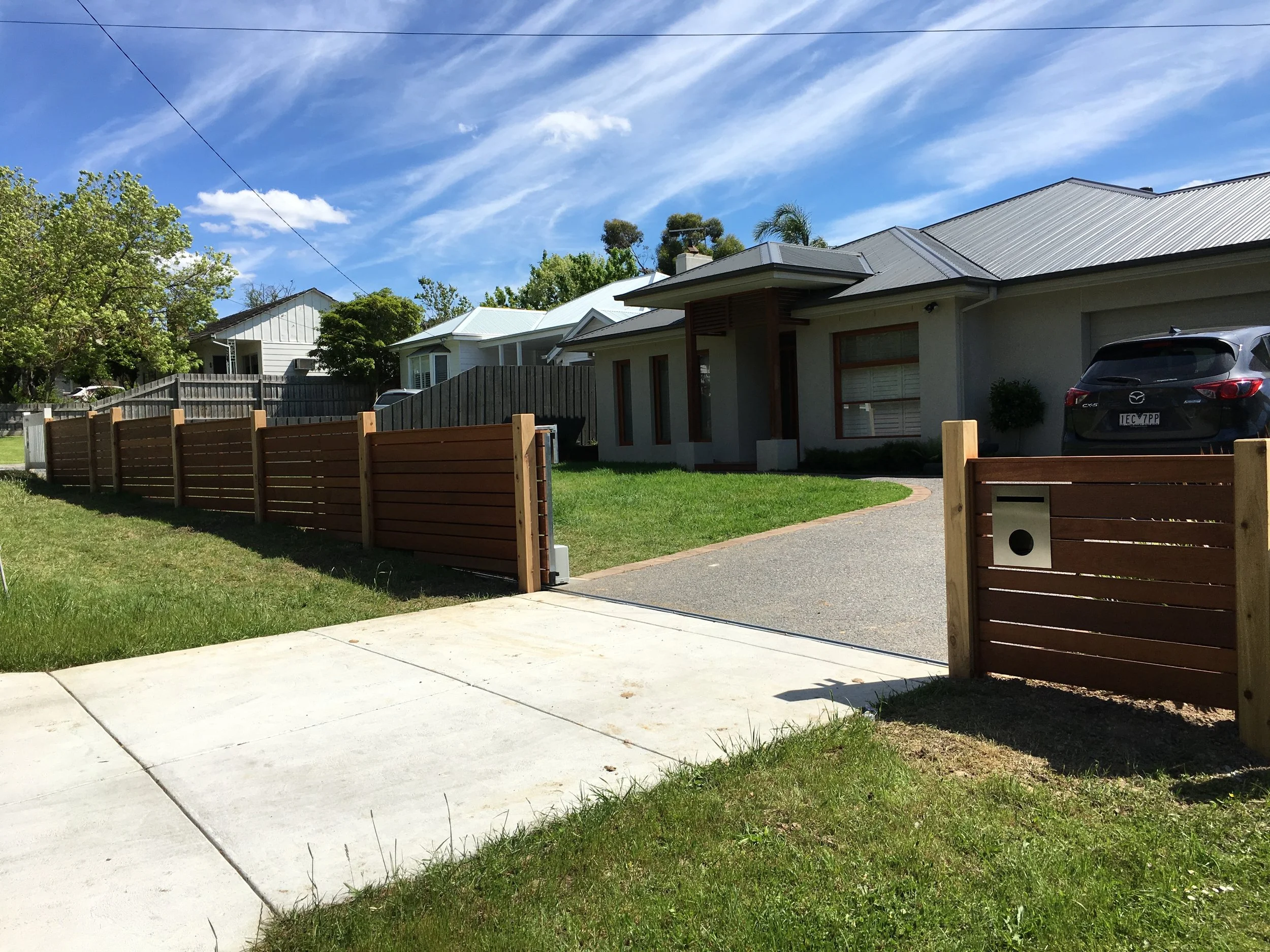 A suburban house with a wooden fence, driveway, and a car parked in the driveway, under a blue sky with clouds.