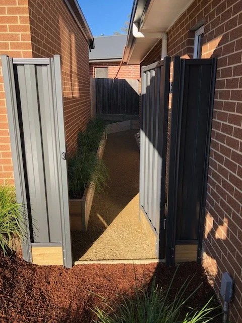 A narrow side yard between two brick houses with a metal fence gate open, showing a concrete walkway and a wooden privacy fence at the end, with some plants on either side.
