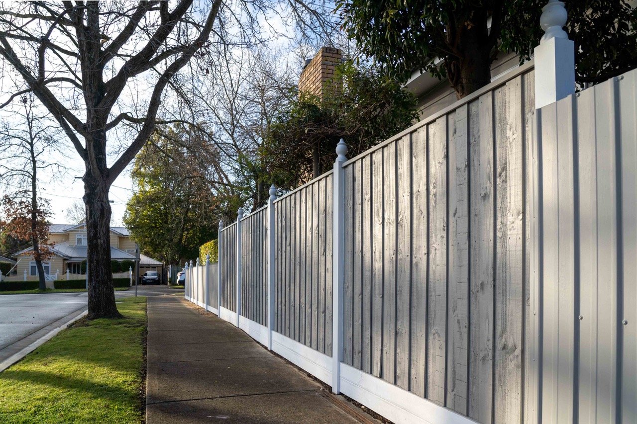 A sidewalk running alongside a residential street with a wooden fence and trees