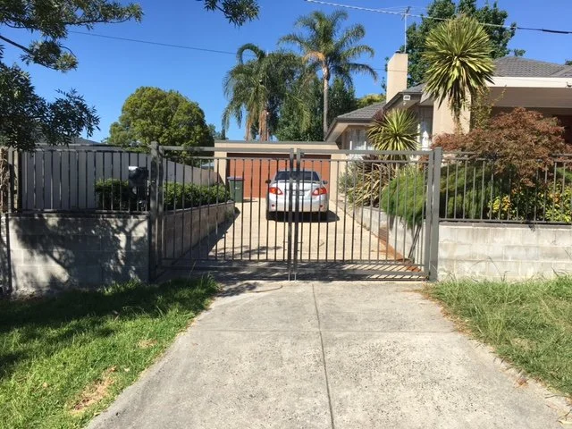 Residential driveway with a metal gate and a parked car, surrounded by plants and trees under a clear blue sky.
