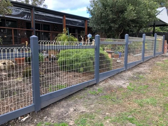 A fenced garden with various plants and decorative items, including statues and a birdhouse, next to a house with a screened porch and trees in the background.