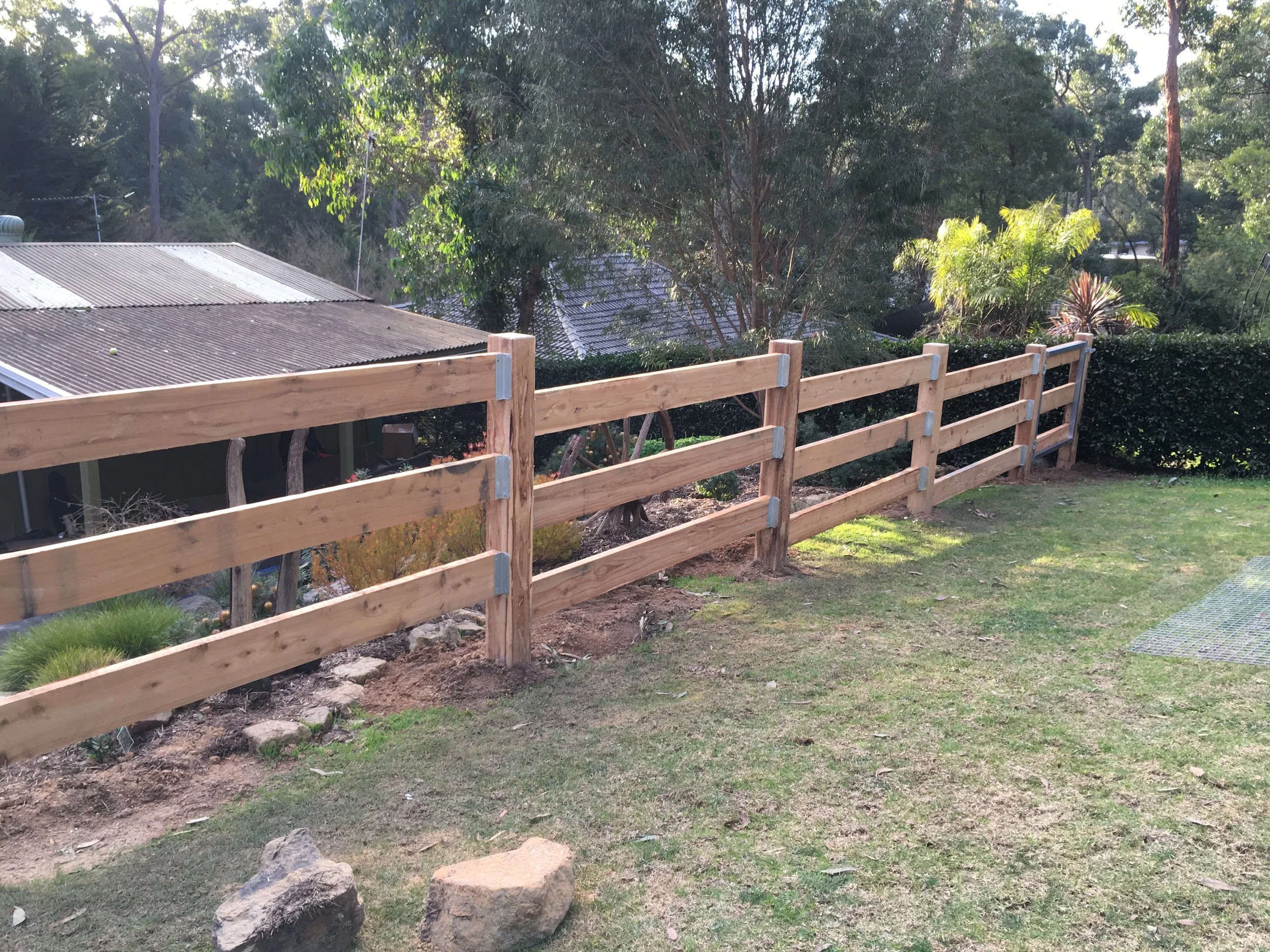 A newly constructed wooden fence with three horizontal planks and metal brackets stands along a grassy backyard, with trees and bushes in the background, and a metal grate on the ground.