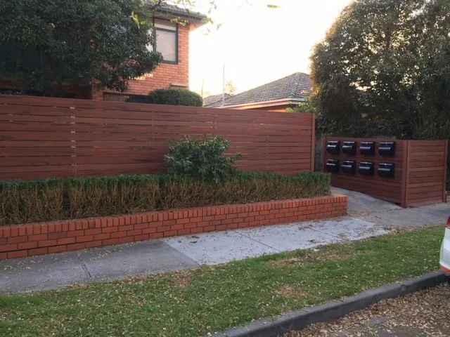 Residential area with wooden fence, mailbox cluster, small bushes, grass, sidewalk, and part of a car visible.