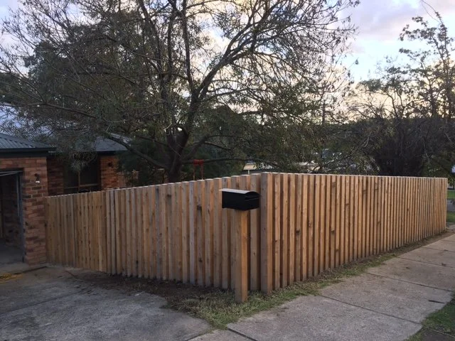 Wooden privacy fence along sidewalk with a mailbox on a post. Behind the fence, trees and houses are visible.