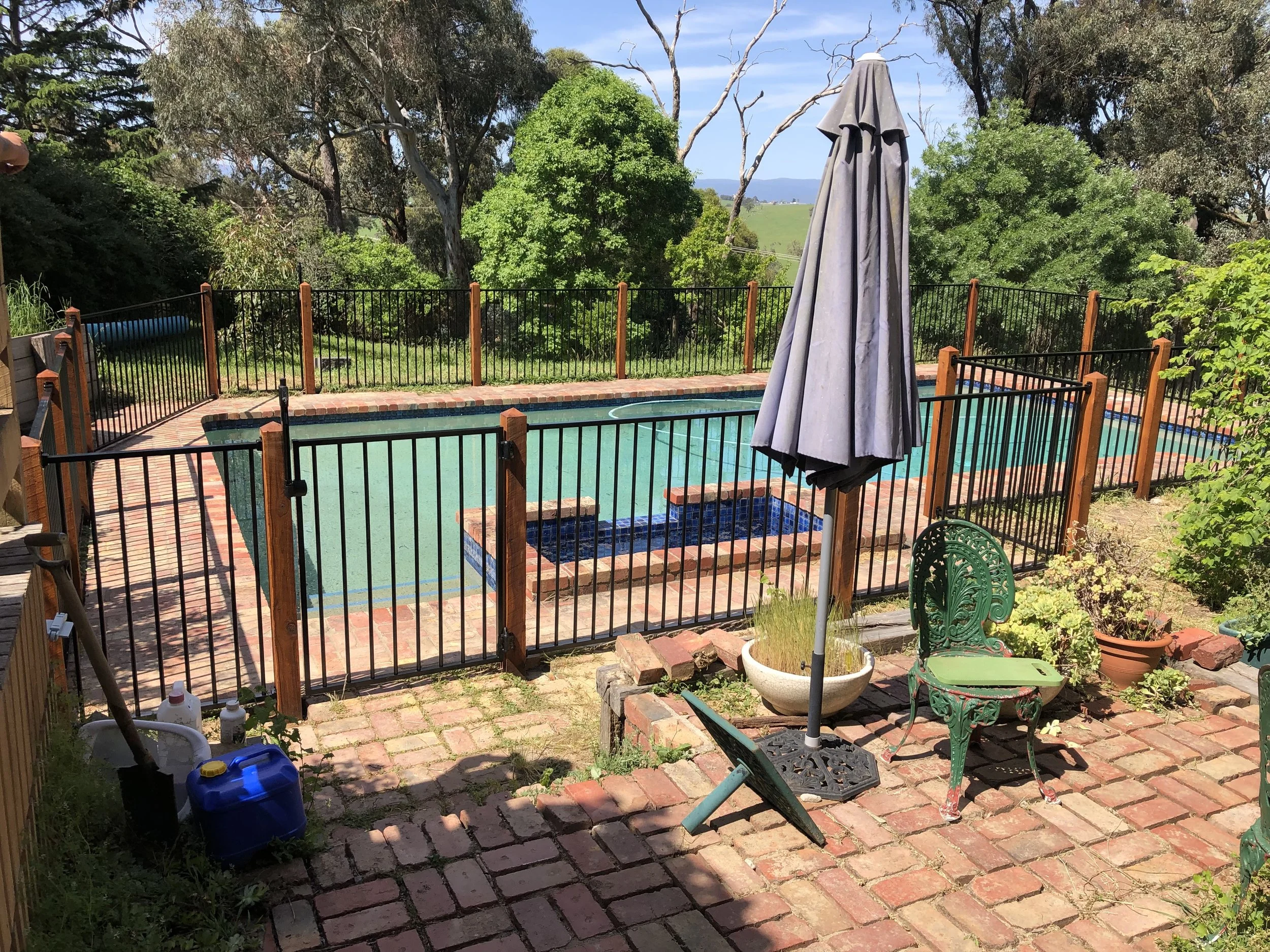 A backyard with a fenced swimming pool, brick patio, outdoor furniture, large umbrella, potted plants, and trees in the background.