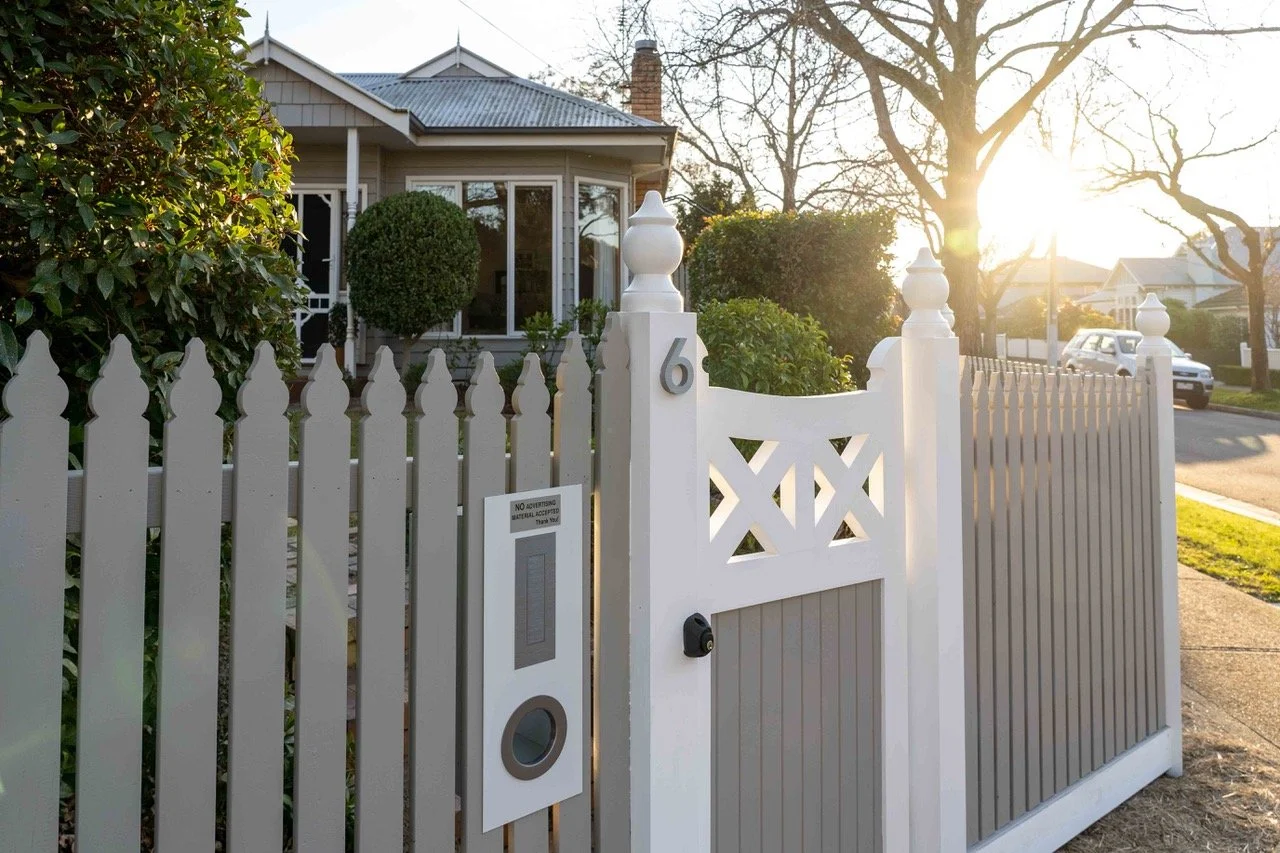 White picket fence gate with house number 6, residential home in background, trees, and sunlight