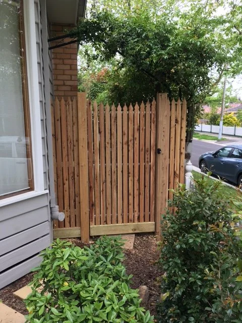Wooden gate in a backyard fence with greenery and parked cars in the background.