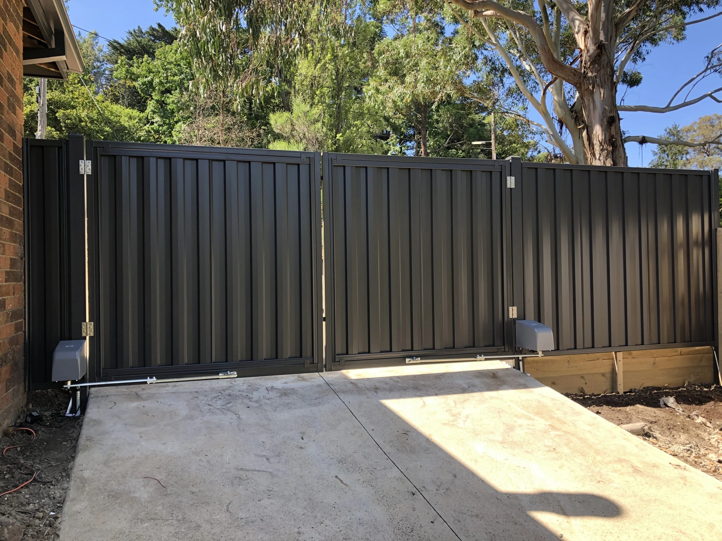 Black metal sliding gate on a concrete driveway, with trees and blue sky in the background.