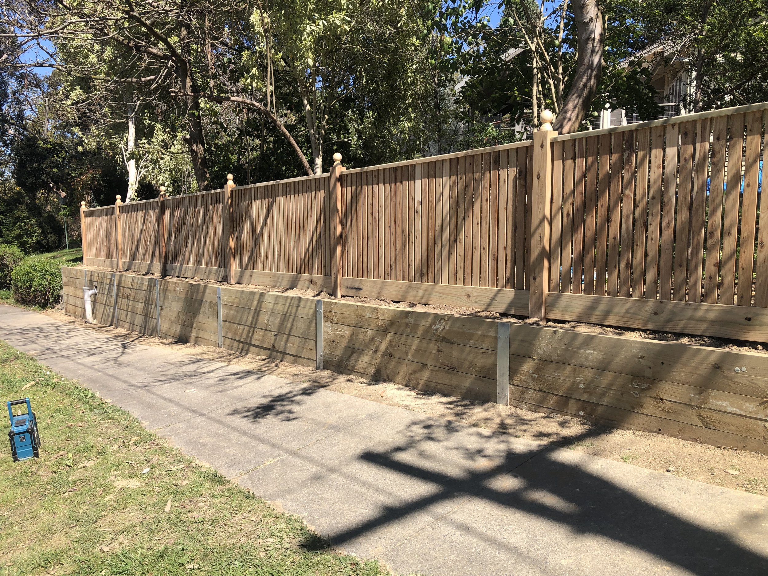 Wooden fence construction along sidewalk with support posts and shaded trees in background.