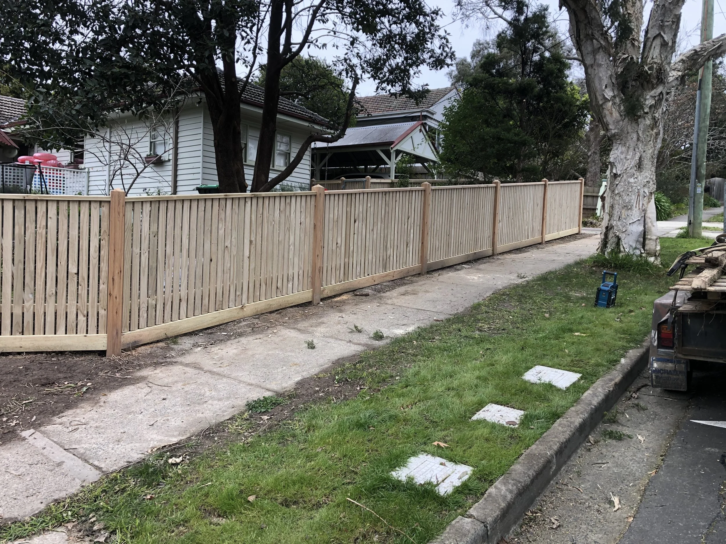 New wooden fence along sidewalk with large tree on right and residential house in background.