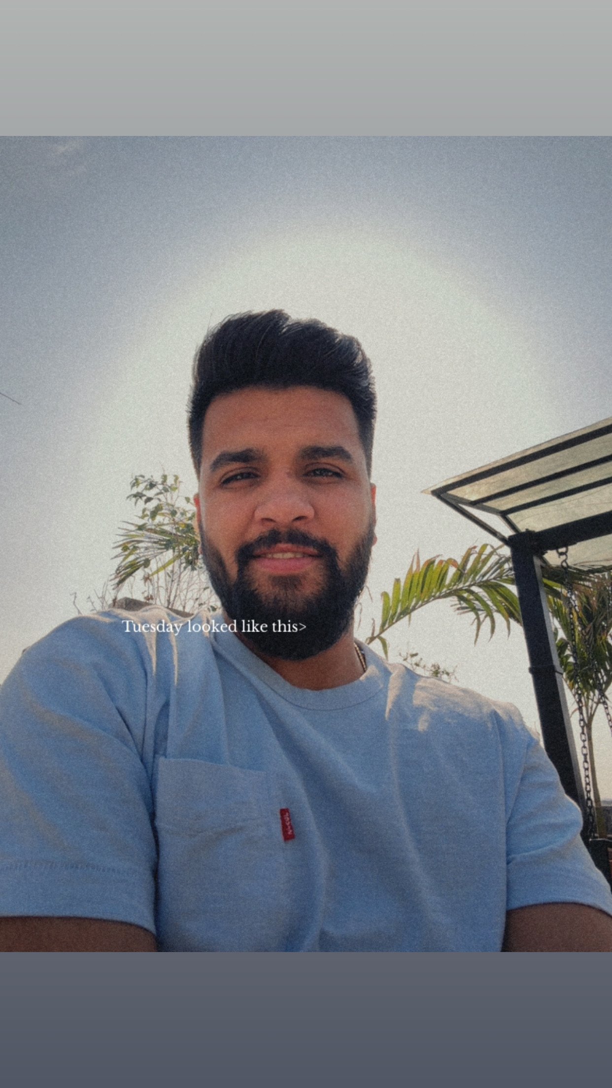 A man with dark hair and a beard taking a selfie outdoors, wearing a light blue shirt with palm trees and a sunny sky in the background.
