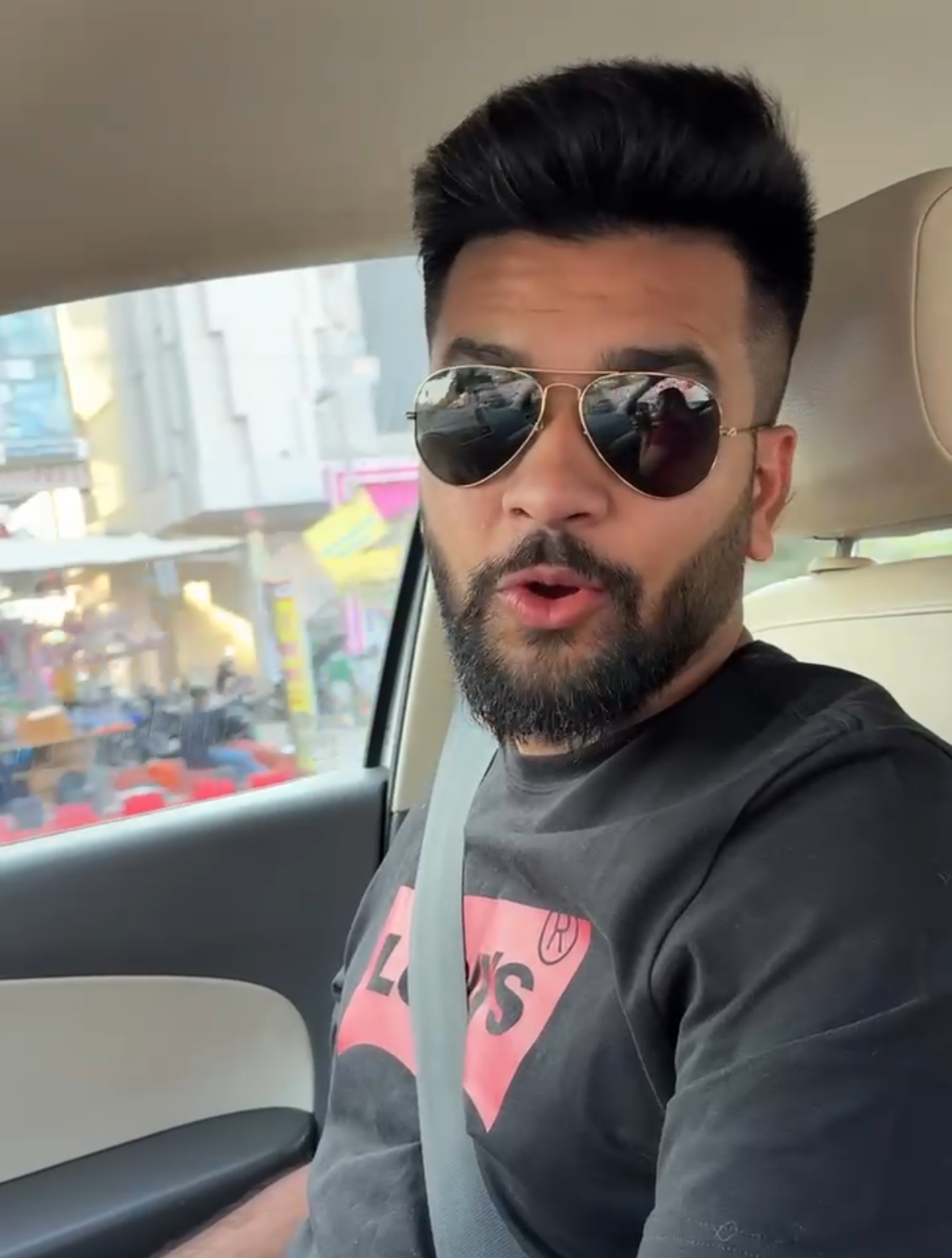 A man with dark hair and a beard wearing sunglasses and a black T-shirt sitting in a car.