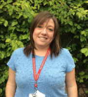 Woman smiling outdoors with green leafy background, wearing a blue top and a red lanyard.