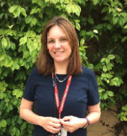 Young woman with brown hair wearing a navy blouse and red lanyard, standing outdoors in front of lush green foliage, smiling at the camera.