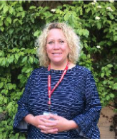 A woman with curly blonde hair standing outdoors in front of green foliage, smiling, wearing a navy blue patterned top and a red lanyard.