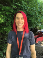 Woman with pink hair standing outdoors next to a car, smiling at the camera.