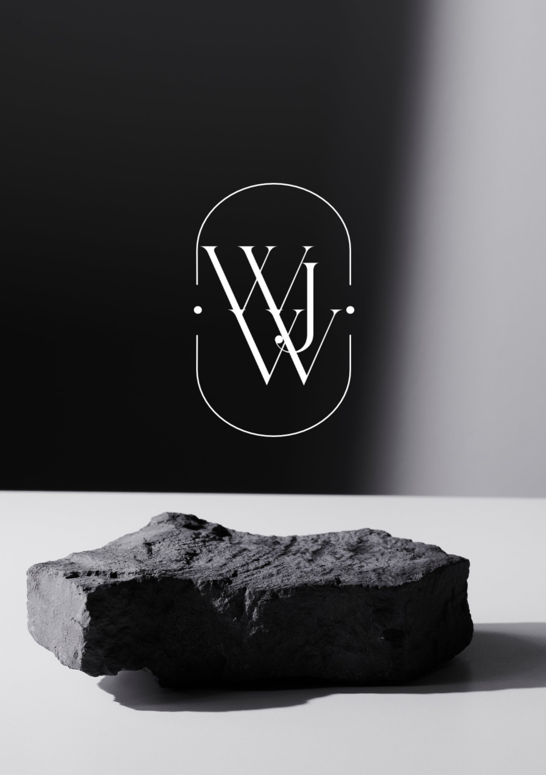 Schwarz-weiß-Fotografie eines Steinblocks auf einer weißen Oberfläche mit einem dunklen Hintergrund, der ein weißes Logo mit den Buchstaben W, J, W zeigt.