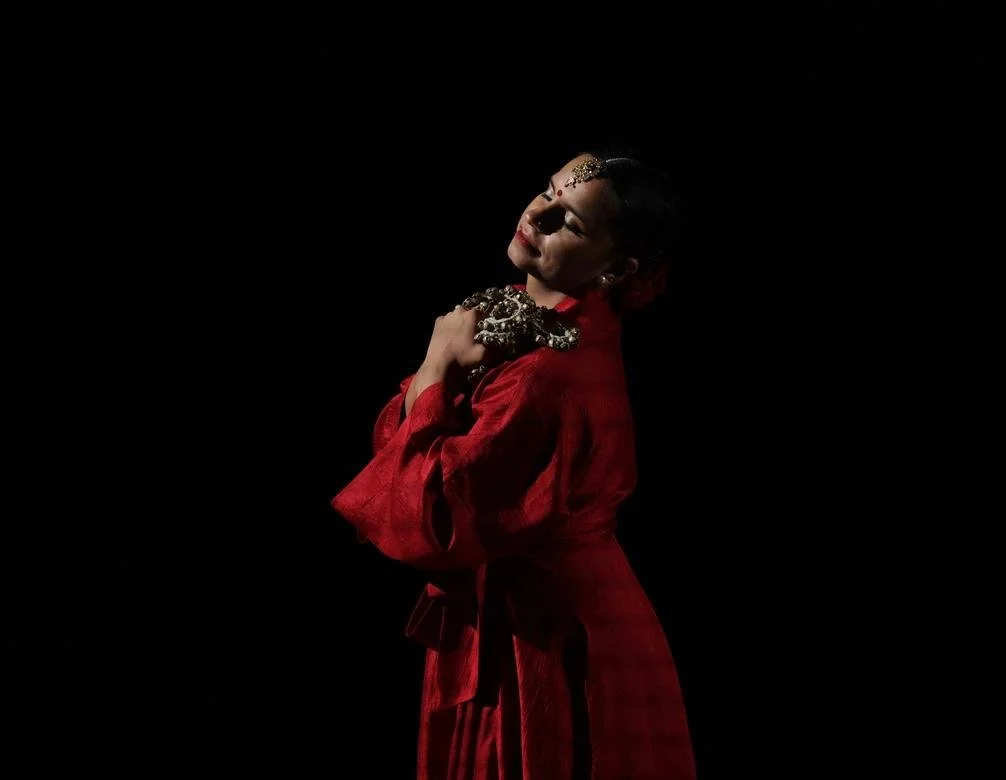 Tarini during performance, posing with ghungroo, wearing red with black backdrop.