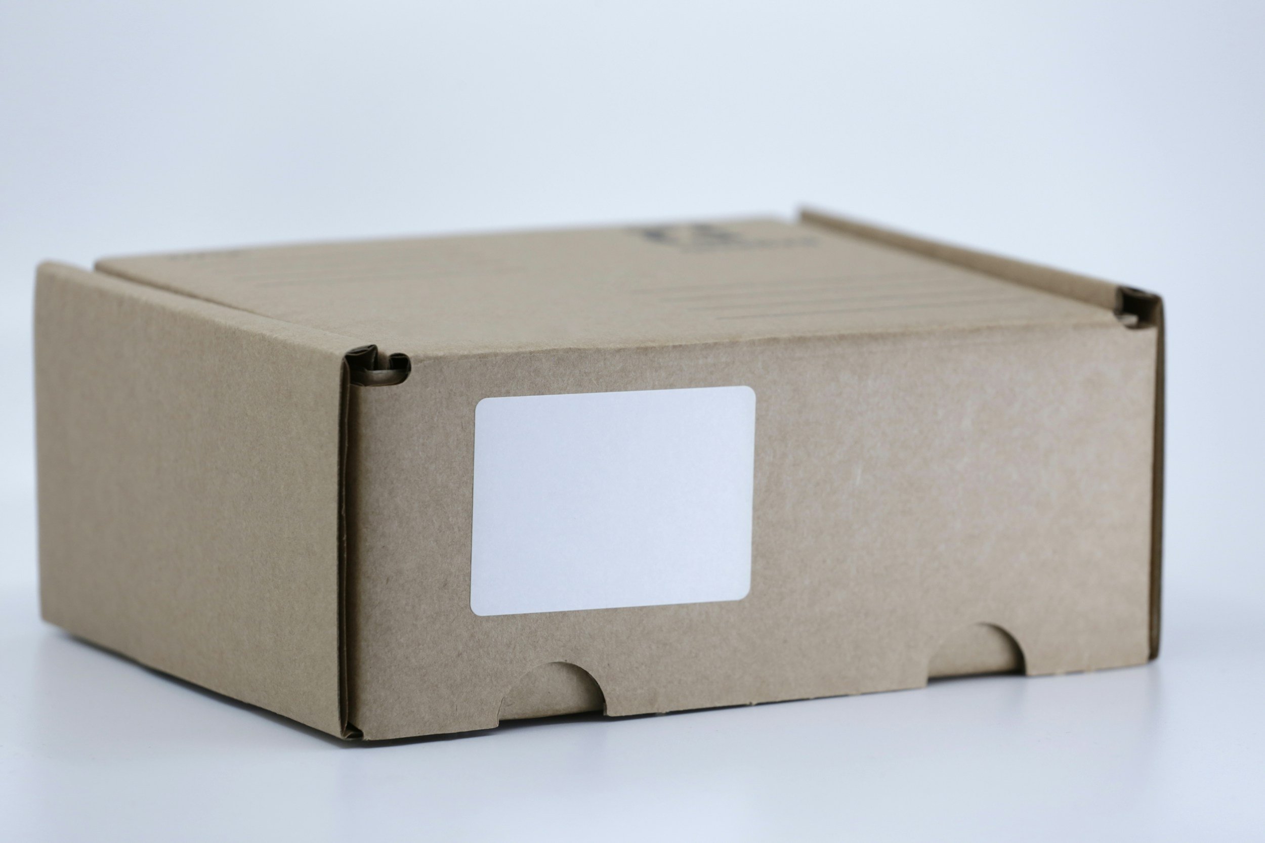 A plain brown cardboard box with a white label on the front, placed on a white surface against a light background.