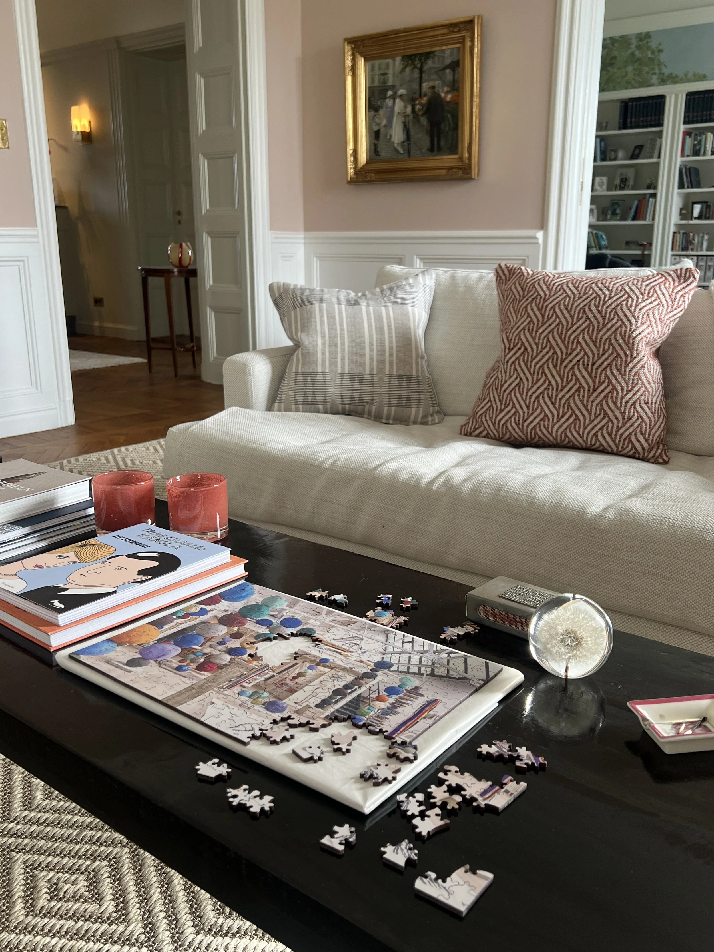 Living room with a beige sofa, decorative pillows, a coffee table with a puzzle, books, candles, and a dandelion in a glass container. Artwork hangs on the wall, a bookshelf is in the background.