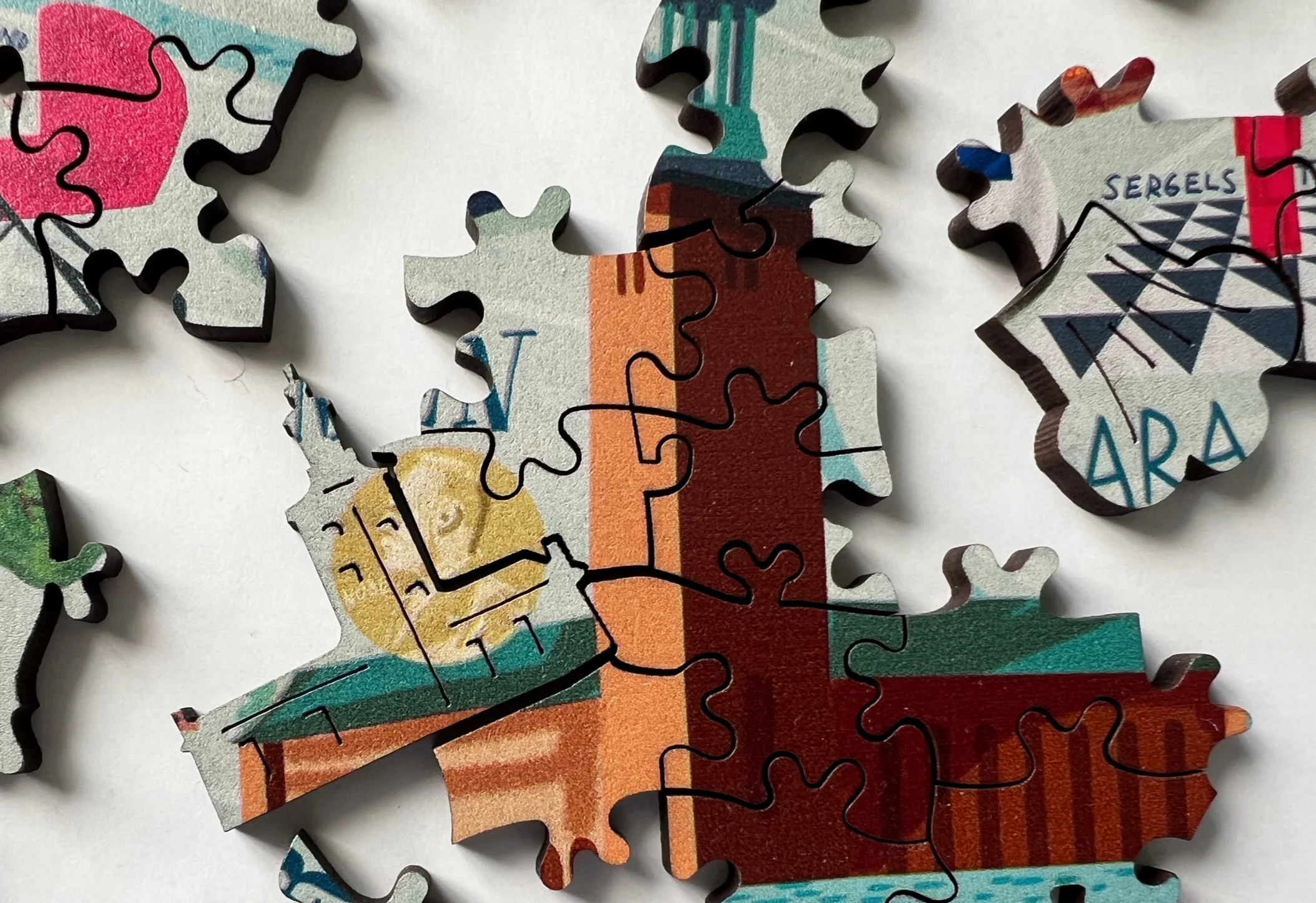Close-up view of a jigsaw puzzle with interlocking pieces, one piece shows a partial illustration of a building with colorful segments and the words 'SERGELS' and 'ARAD' on some pieces.