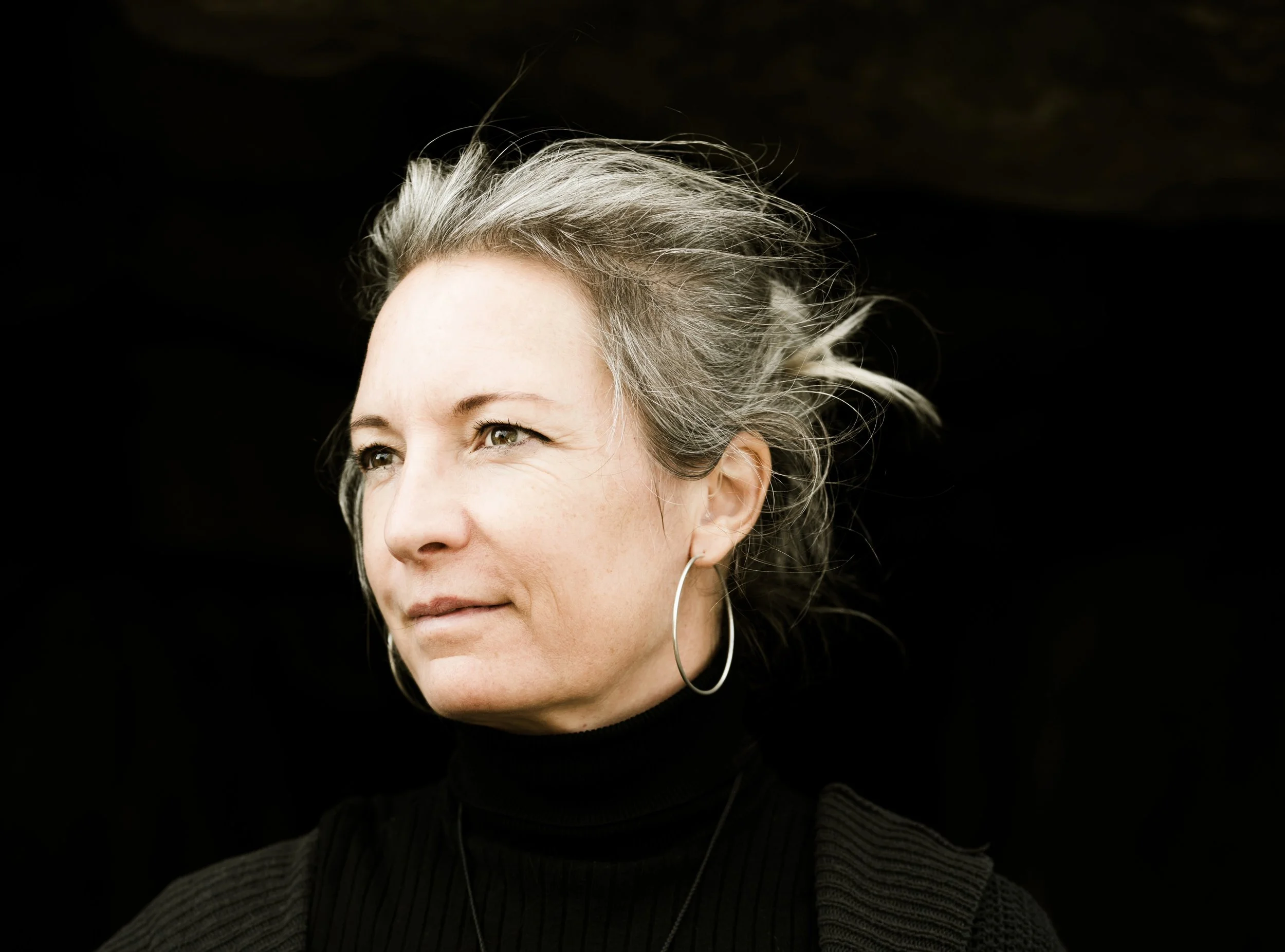 Nahaufnahme einer Frau mit grauem Haar und großen Ohrringen, die nach rechts schaut, vor dunklem Hintergrund.